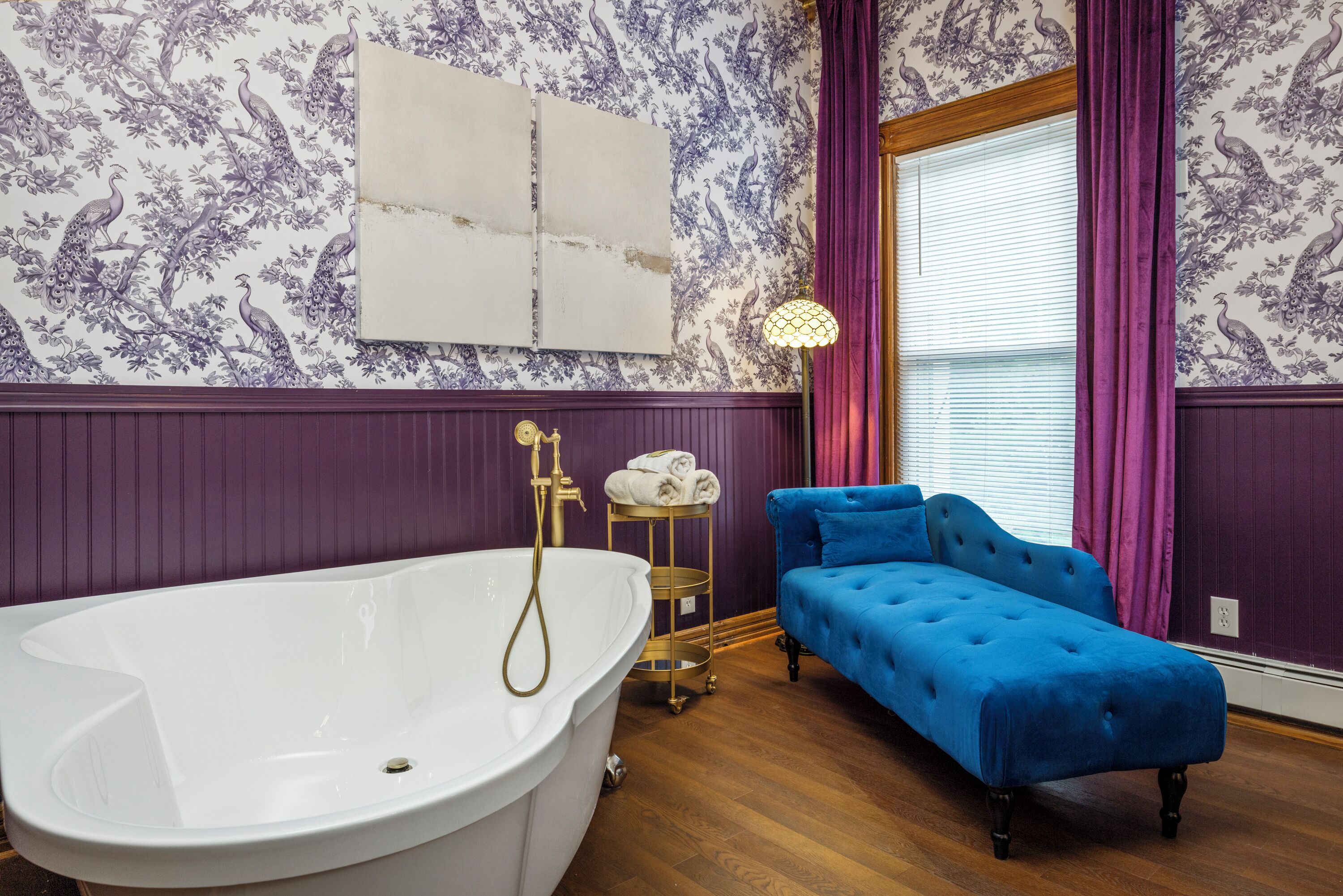 A bathroom with a freestanding bathtub, a blue sofa, and a window with purple curtains.