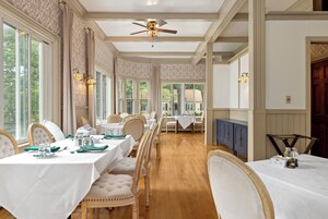 Une salle à manger avec plusieurs tables dressées pour le repas, de grandes fenêtres et un ventilateur de plafond.
