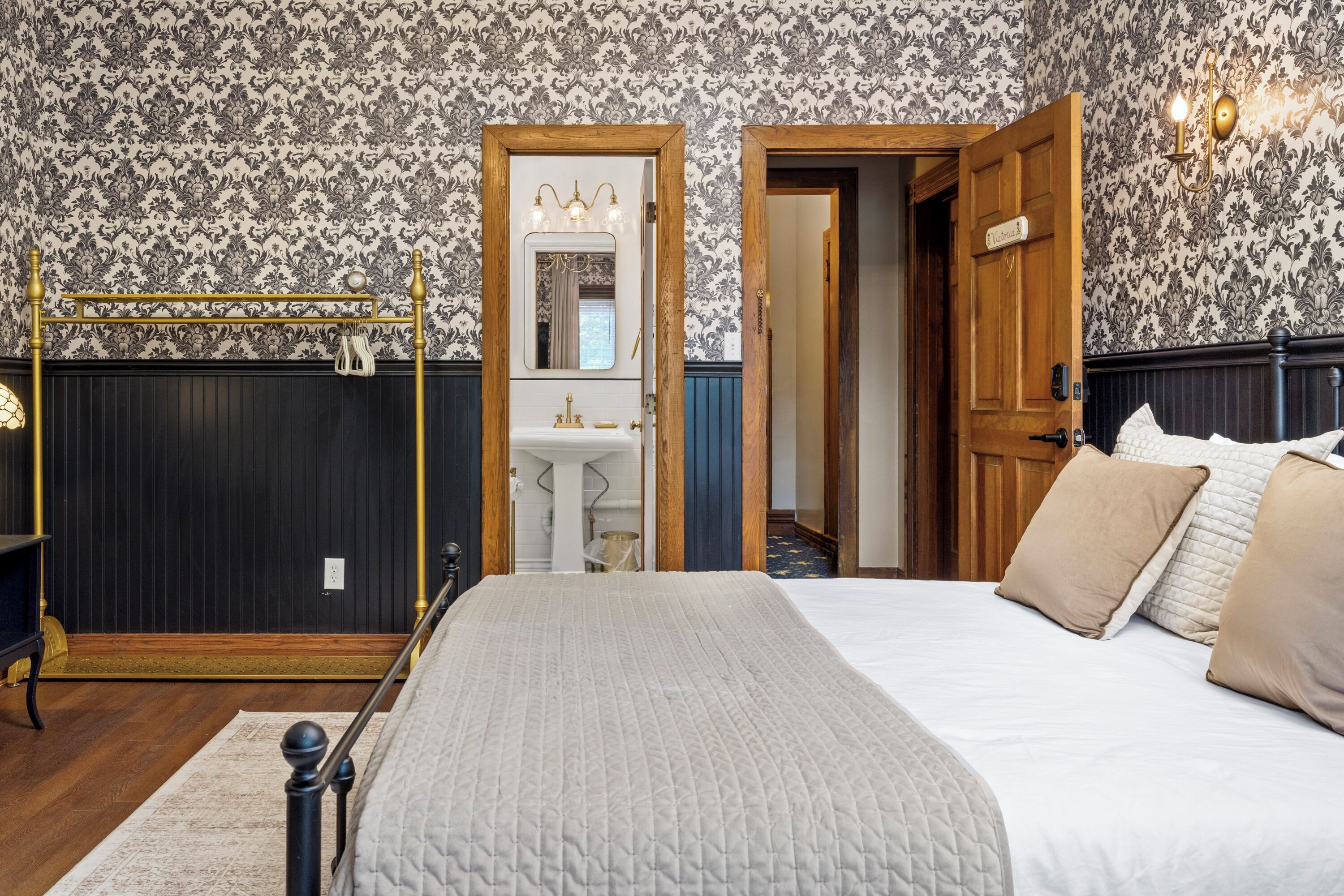 A bedroom with a bed, a wooden door, a mirror, and a wall with patterned wallpaper.