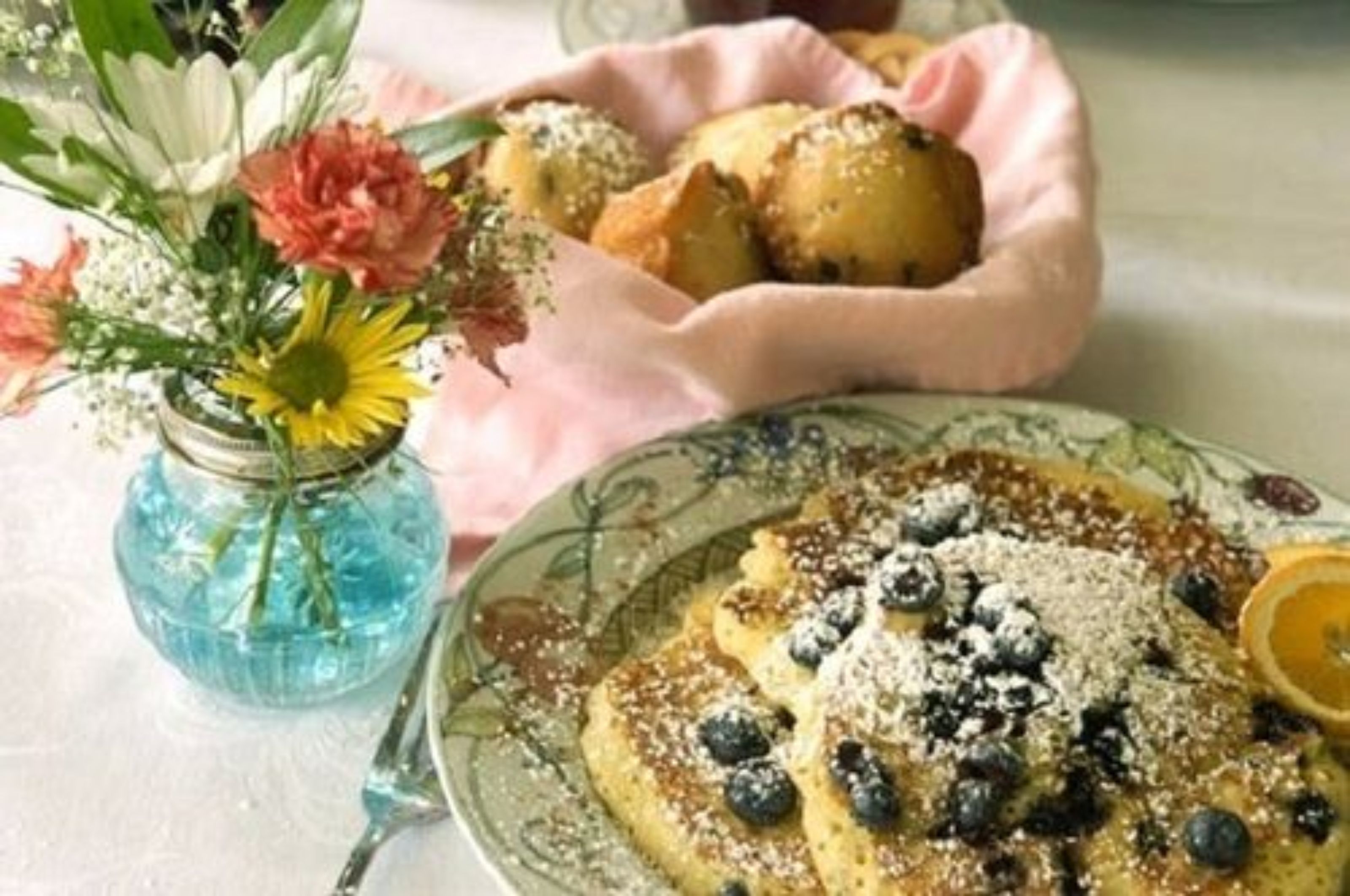 A breakfast setting with pancakes topped with blueberries and powdered sugar, a vase with flowers, and a basket of pastries.