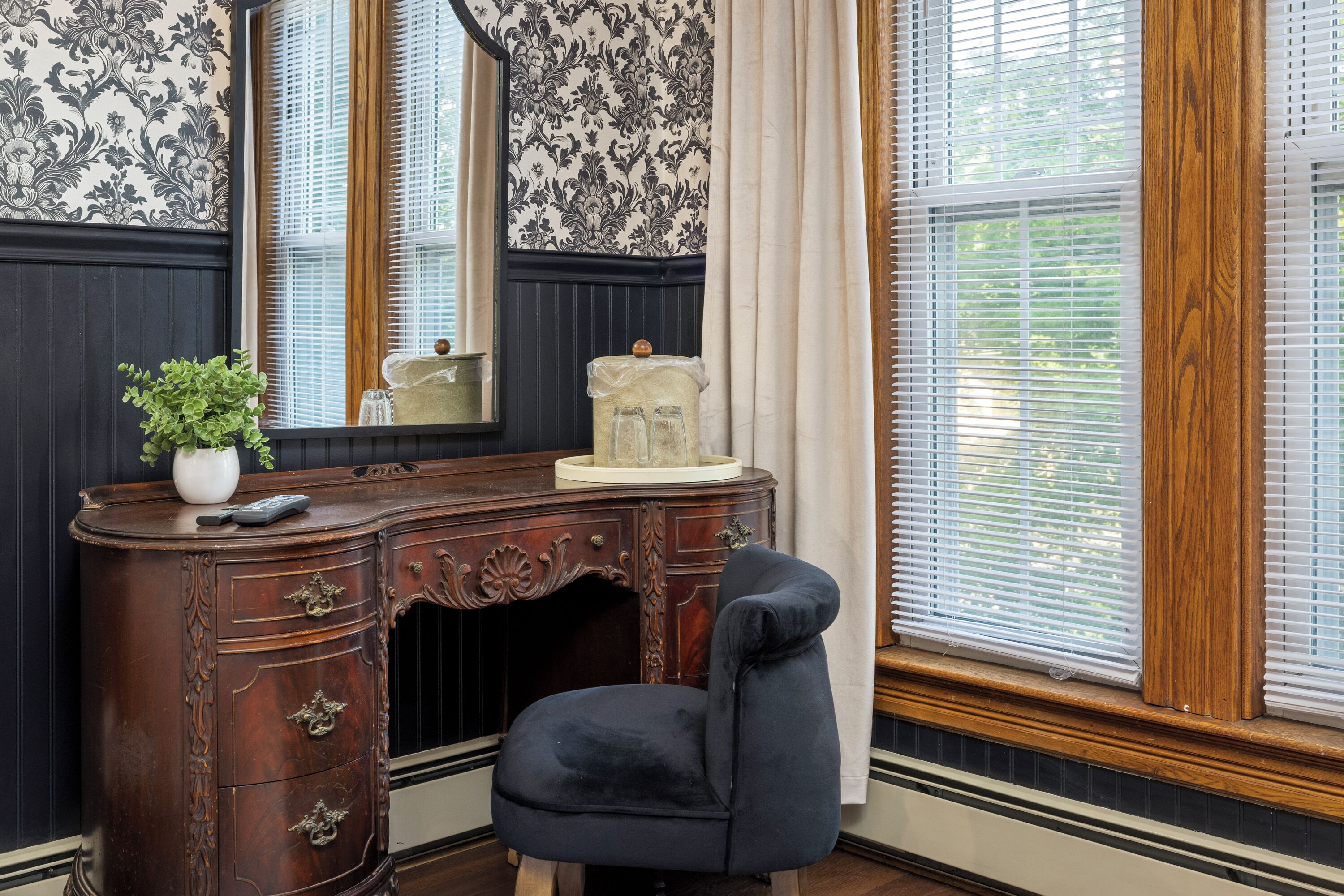 A room with a dark wooden desk, a chair, a potted plant, and a window with blinds.