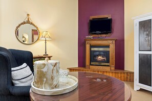 A living room with a fireplace, a television mounted above it, a round wooden coffee table, and a blue armchair with a decorative pillow. - Rockwell Falls Inn and Wine Bar (Lake Luzerne)