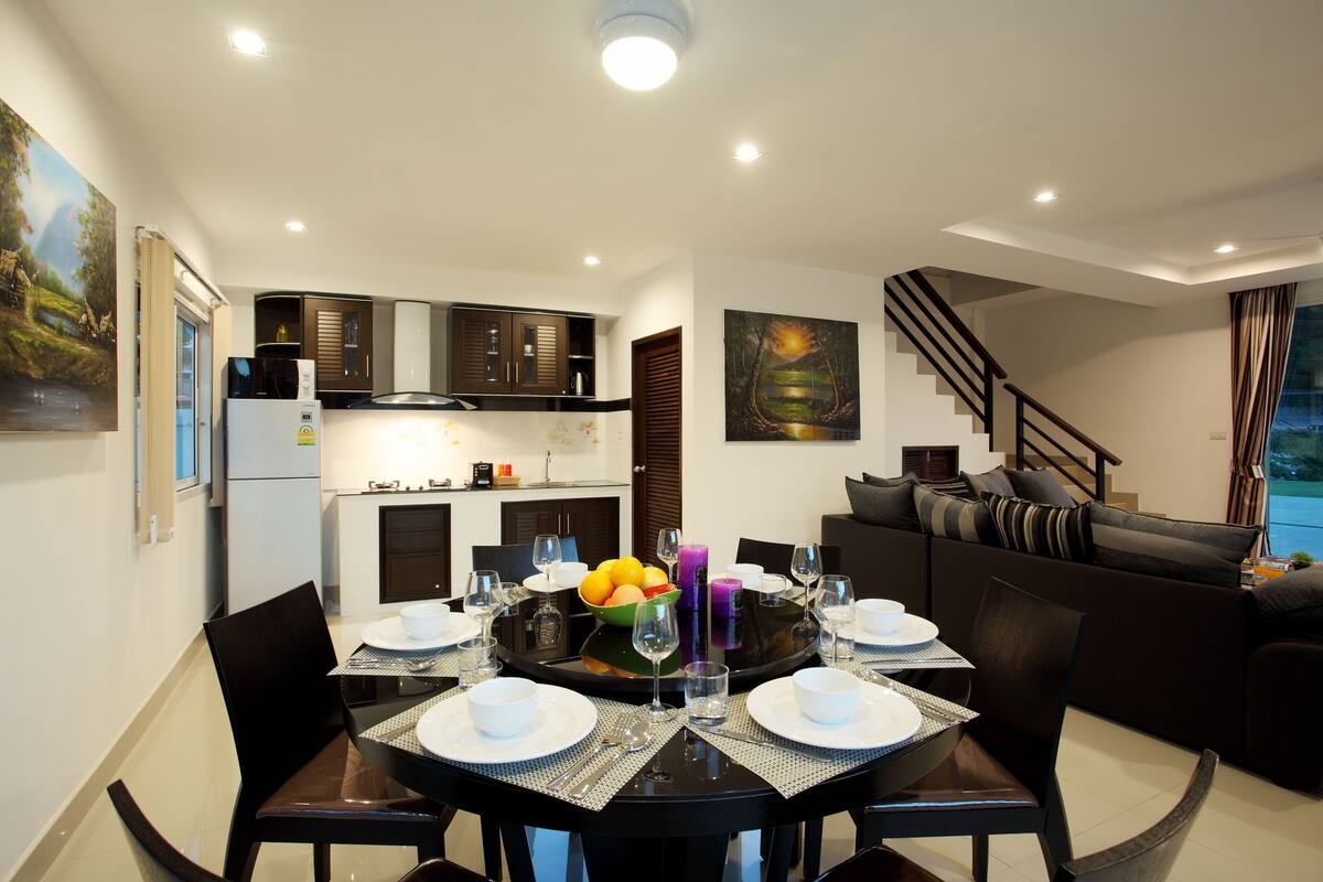Een moderne eethoek met een ronde tafel voor vier personen, een keuken op de achtergrond en een trap naar een woongedeelte.