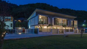 Een modern huis met twee verdiepingen, een plat dak, grote ramen en een goed verlichte voortuin.