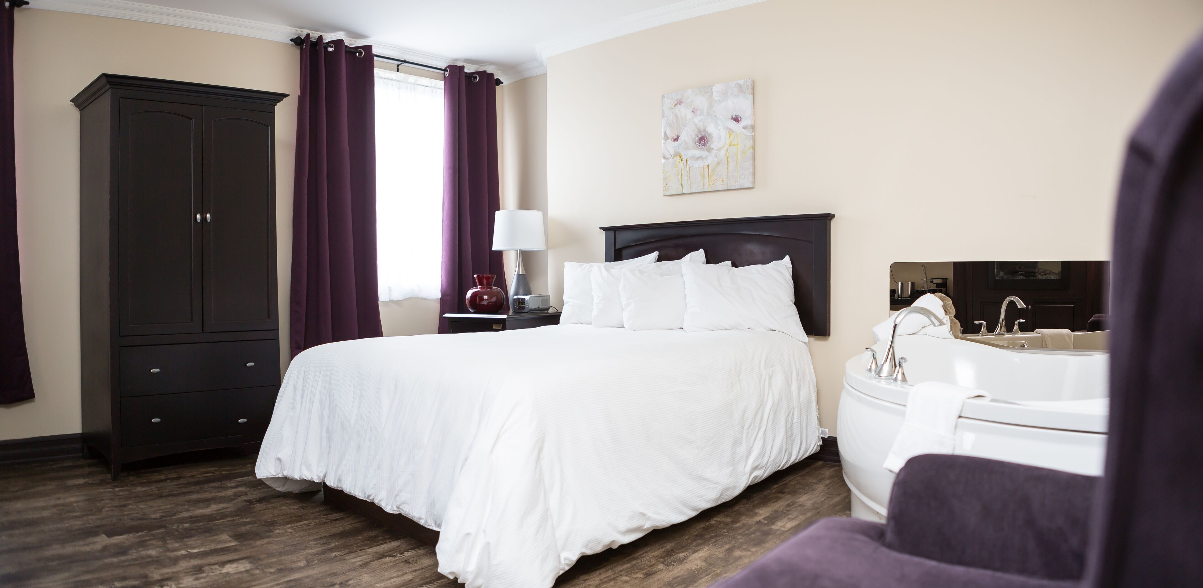 A hotel room with a large bed, a wardrobe, a nightstand with a lamp, a painting on the wall, and a bathroom with a bathtub and sink.