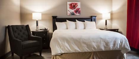 A hotel room with a large bed, two bedside tables with lamps, a dark armchair, and a floral artwork on the wall.