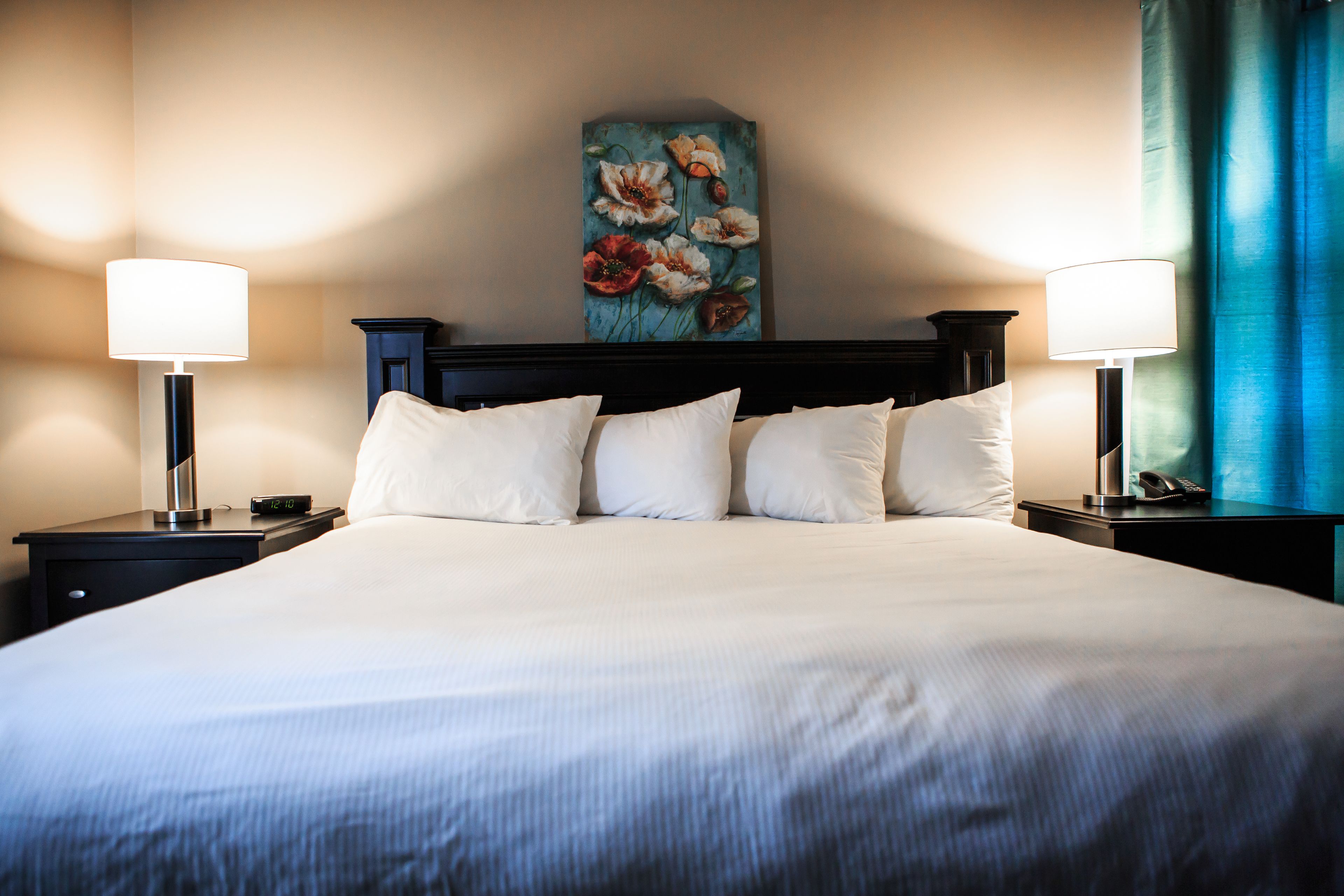A neatly made bed with white linens and a dark headboard. There are two bedside tables, each with a lamp and a clock. A painting of flowers hangs on the wall above the bed.