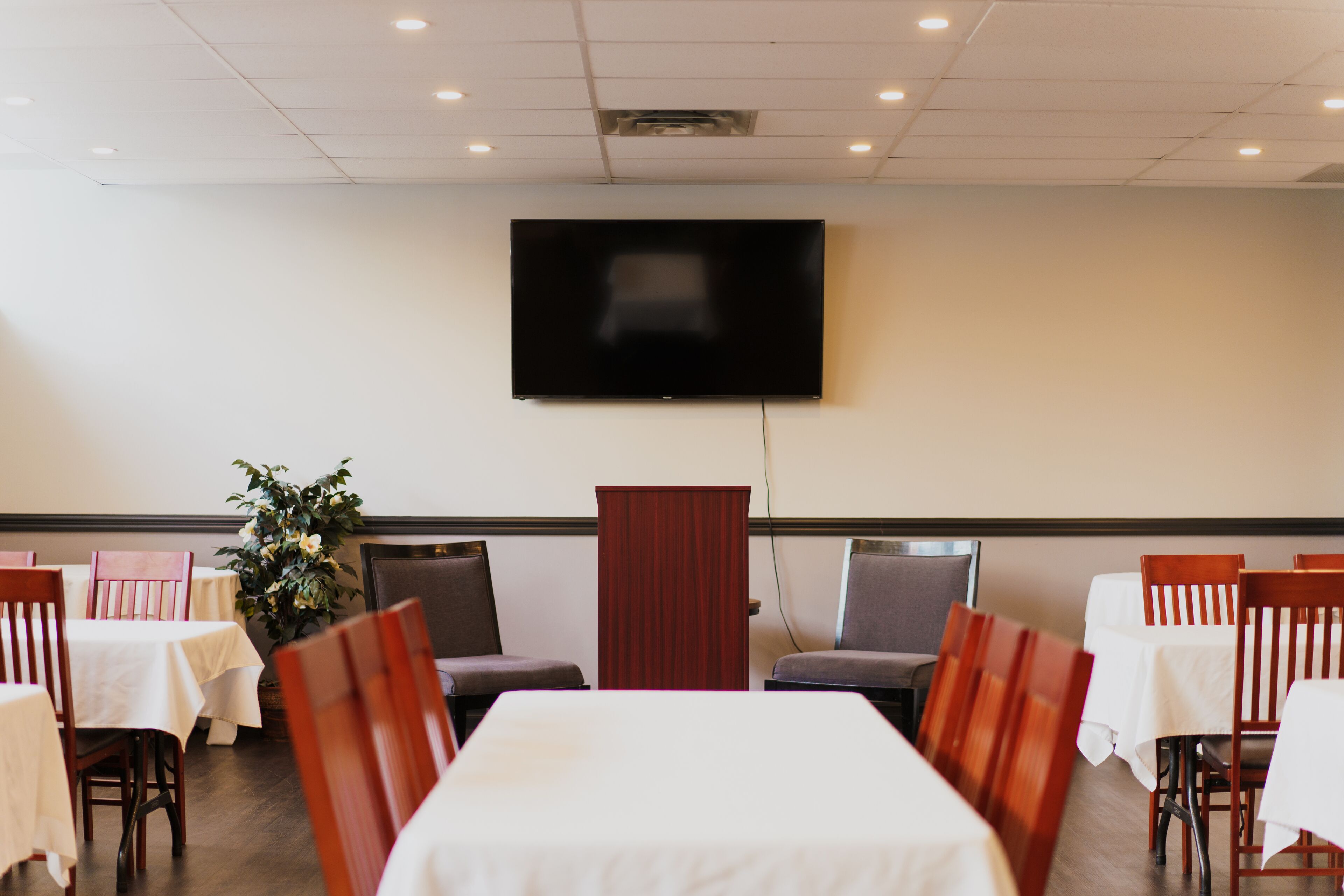 A conference room with a large flat-screen TV mounted on the wall, a podium, and rows of tables and chairs.