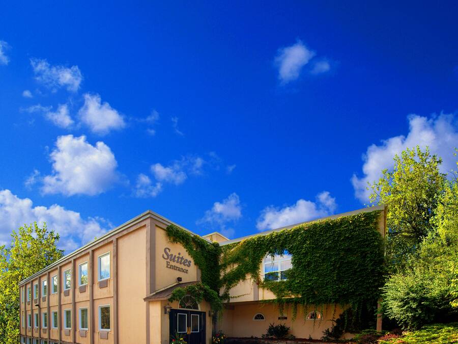 Entrada de hotel con un letrero que dice "Suites Entrance", un camino pavimentado y un cielo azul despejado con nubes.