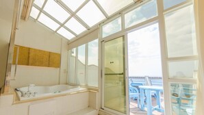 Une salle de bain dotée d’une grande baignoire, d’une douche à parois de verre et offrant une vue sur la mer grâce à de grandes fenêtres.