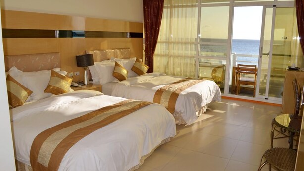 Une chambre d’hôtel avec deux lits, un bureau, une chaise et une vue sur la mer, visible à travers de grandes portes vitrées.