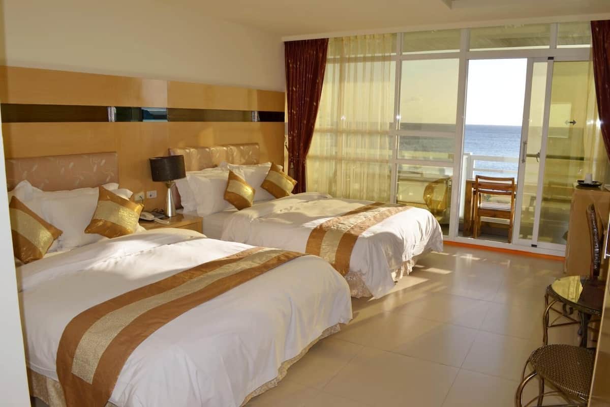 A hotel room with two beds, a desk, a chair, and a view of the sea through large glass doors.