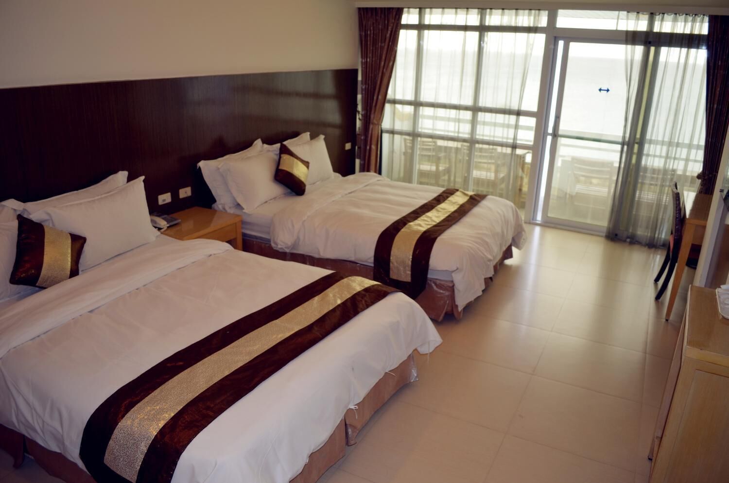 Habitación de hotel con dos camas, cabecera de madera, un ventanal grande con cortinas y un escritorio de madera.