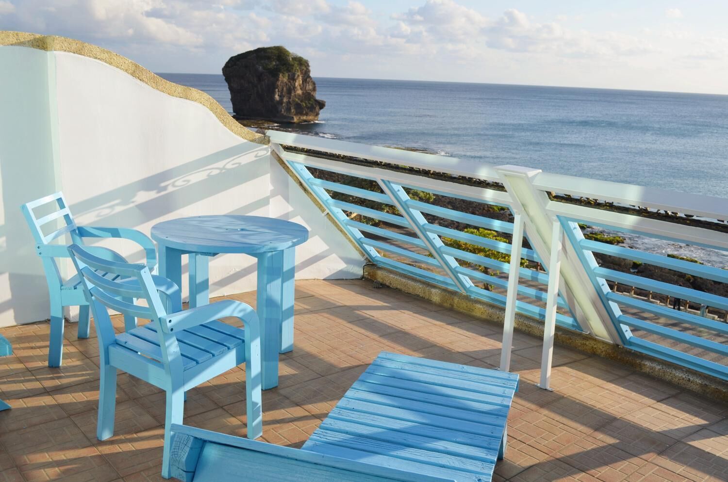 A terrace with blue furniture, a view of the sea, and a rocky outcrop.