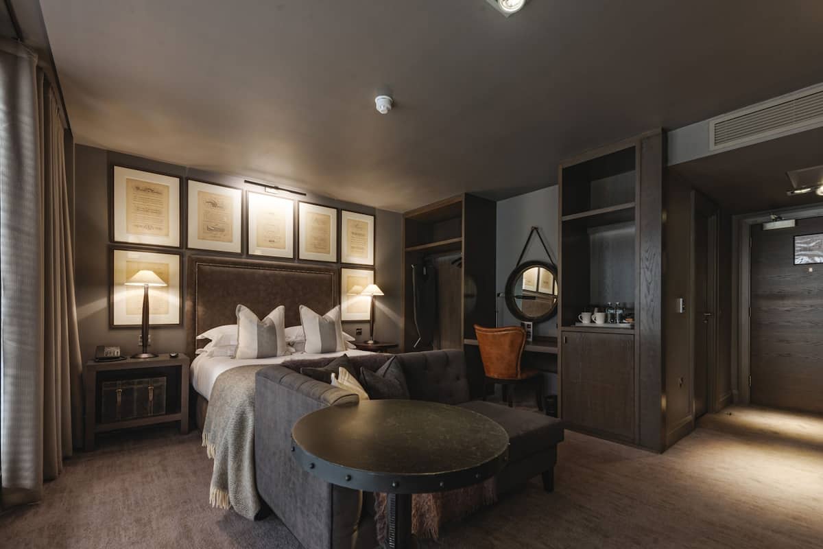 Habitación de hotel moderna con cama, sofá, mesa redonda y escritorio. En la pared hay certificados enmarcados, un espejo redondo y una pequeña mesita de noche con lámpara.