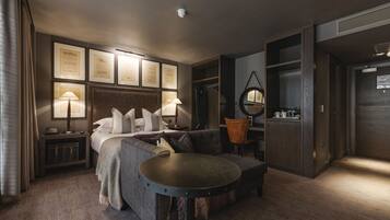 Habitación de hotel moderna con cama, sofá, mesa redonda y escritorio. En la pared hay certificados enmarcados, un espejo redondo y una pequeña mesita de noche con lámpara.