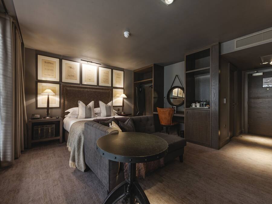 Habitación de hotel moderna con cama, sofá, mesa redonda y escritorio. En la pared hay certificados enmarcados, un espejo redondo y una pequeña mesita de noche con lámpara.