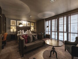 Una habitación de hotel moderna con una cama grande, un sofá, una mesa redonda y vistas de la ciudad a través de amplios ventanales.