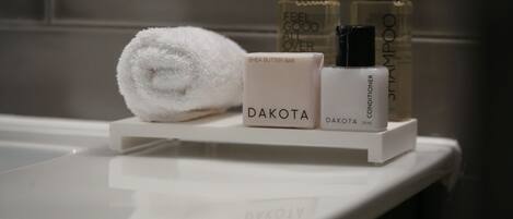 Una encimera de baño con una toalla blanca enrollada, una pastilla de Dakota de manteca de shea y dos botellas de acondicionador y champú Dakota.