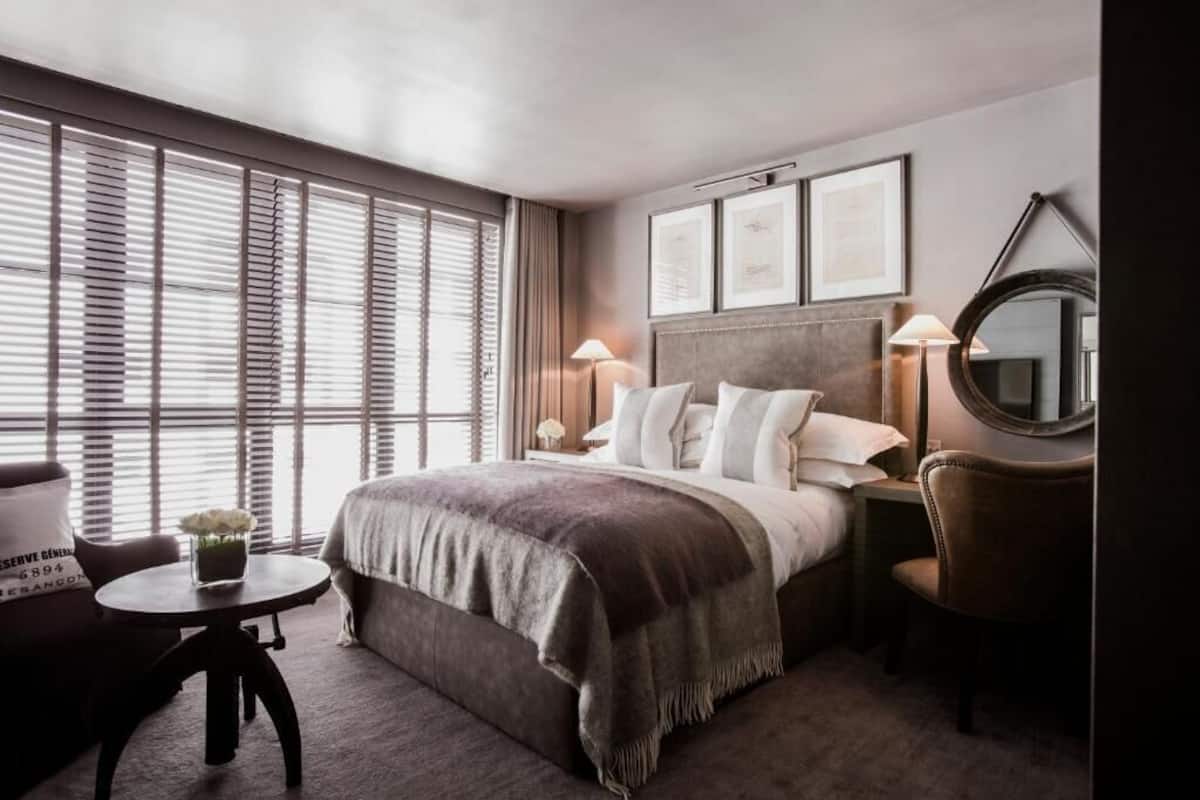 Habitación de hotel con cama, lámparas de noche, un espejo redondo, una silla, una mesita con un jarrón y una ventana con persianas.