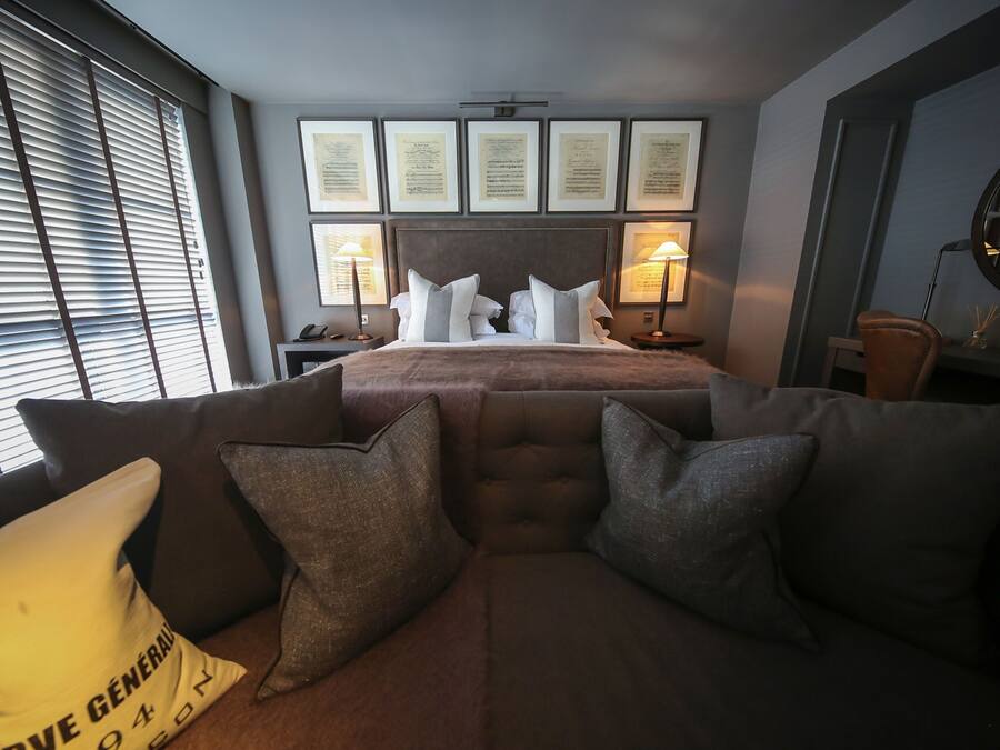 Ein modernes Hotelzimmer mit einem großen Bett, einer Couch mit vielen Kissen, gerahmten Zertifikaten an der Wand und einem Fenster mit Jalousien.