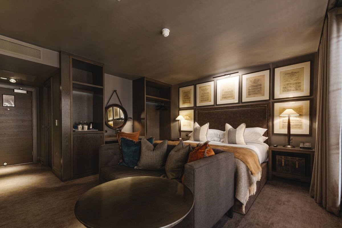 Habitación de hotel con una cama, una mesa redonda, un sofá, un espejo, una lámpara y diplomas enmarcados en la pared.