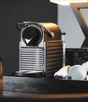 Una máquina de café Nespresso con diseño negro y cromado, sobre una encimera negra con una lámpara al fondo.