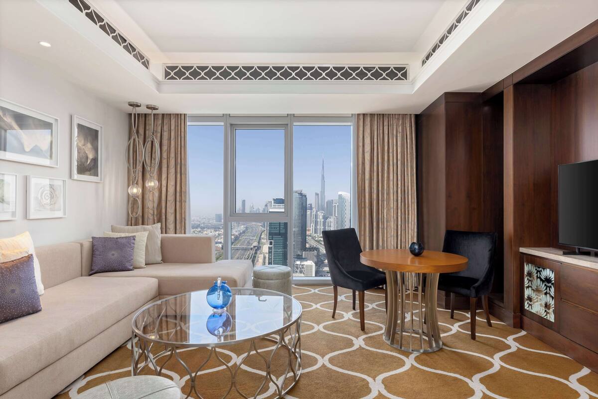 Habitación de hotel moderna con un amplio ventanal que ofrece vistas a la ciudad, un sofá, una mesa de centro y un televisor.