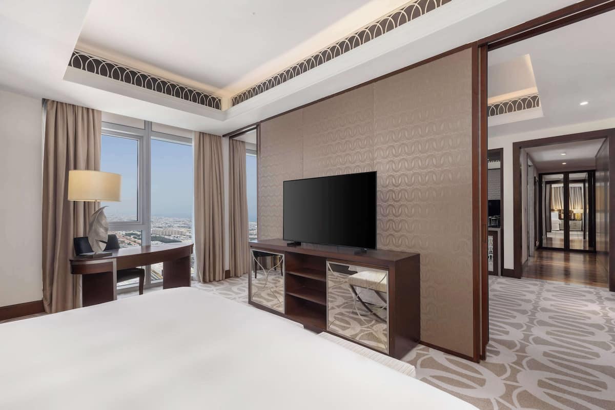 Habitación de hotel con una cama grande, un televisor sobre un soporte de madera, un escritorio con lámpara y vistas a la ciudad a través de amplias ventanas.