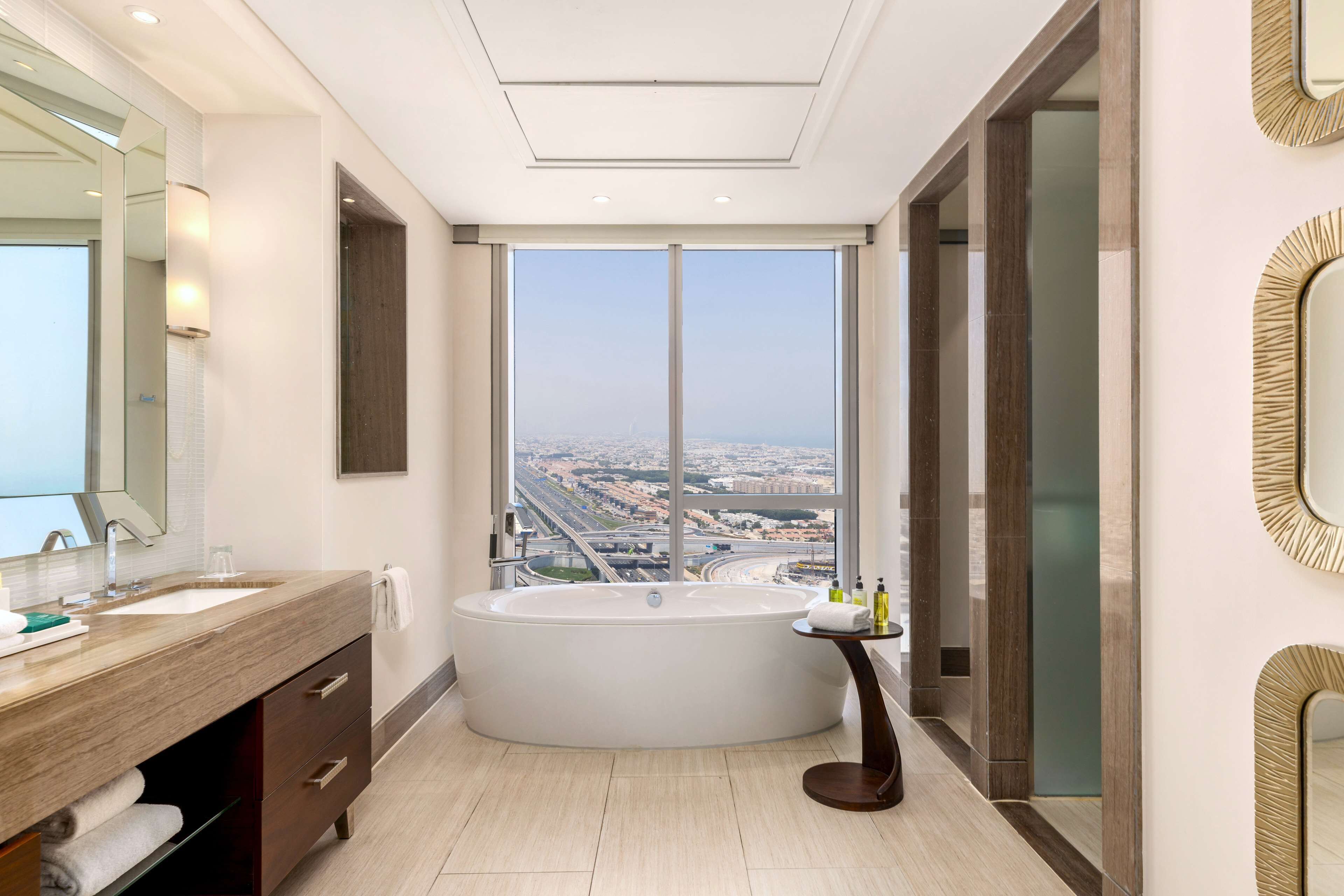 Une salle de bain moderne dotée d’une grande baignoire, d’un meuble-lavabo avec un miroir et offrant une vue sur le paysage urbain grâce à une grande fenêtre.