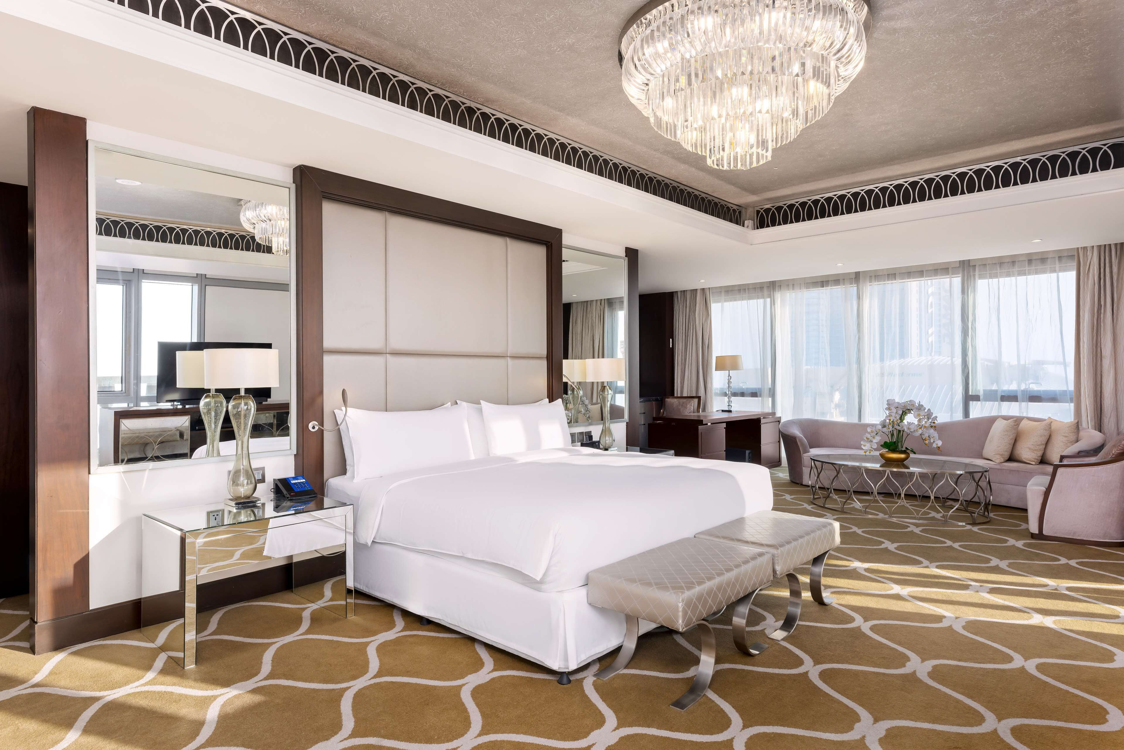 A spacious hotel room with a large bed, a sitting area with a sofa and armchairs, a dining table, and a large window offering a city view.