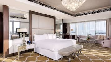 A spacious hotel room with a large bed, a sitting area with a sofa and armchairs, a dining table, and a large window offering a city view.