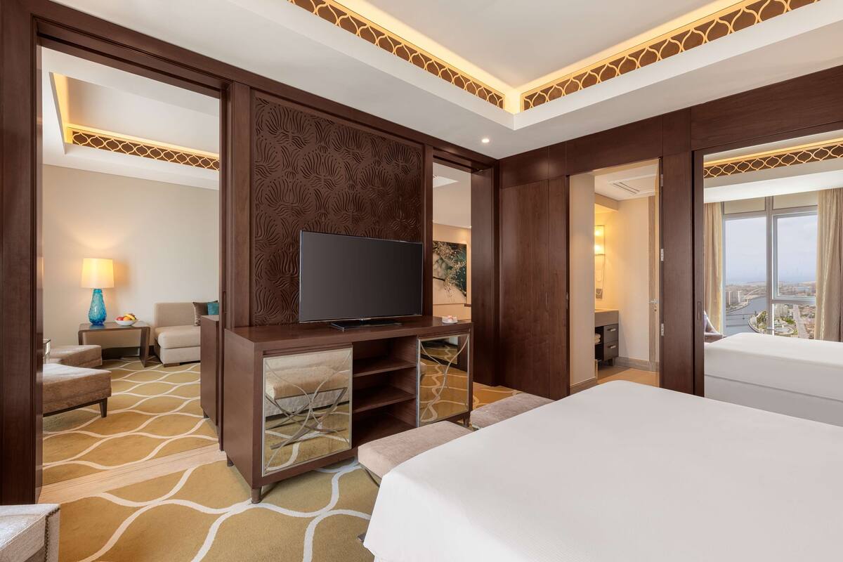 Habitación de hotel con una cama grande, un televisor sobre un mueble de madera, un rincón de descanso con una lámpara y vistas a la ciudad desde la ventana.