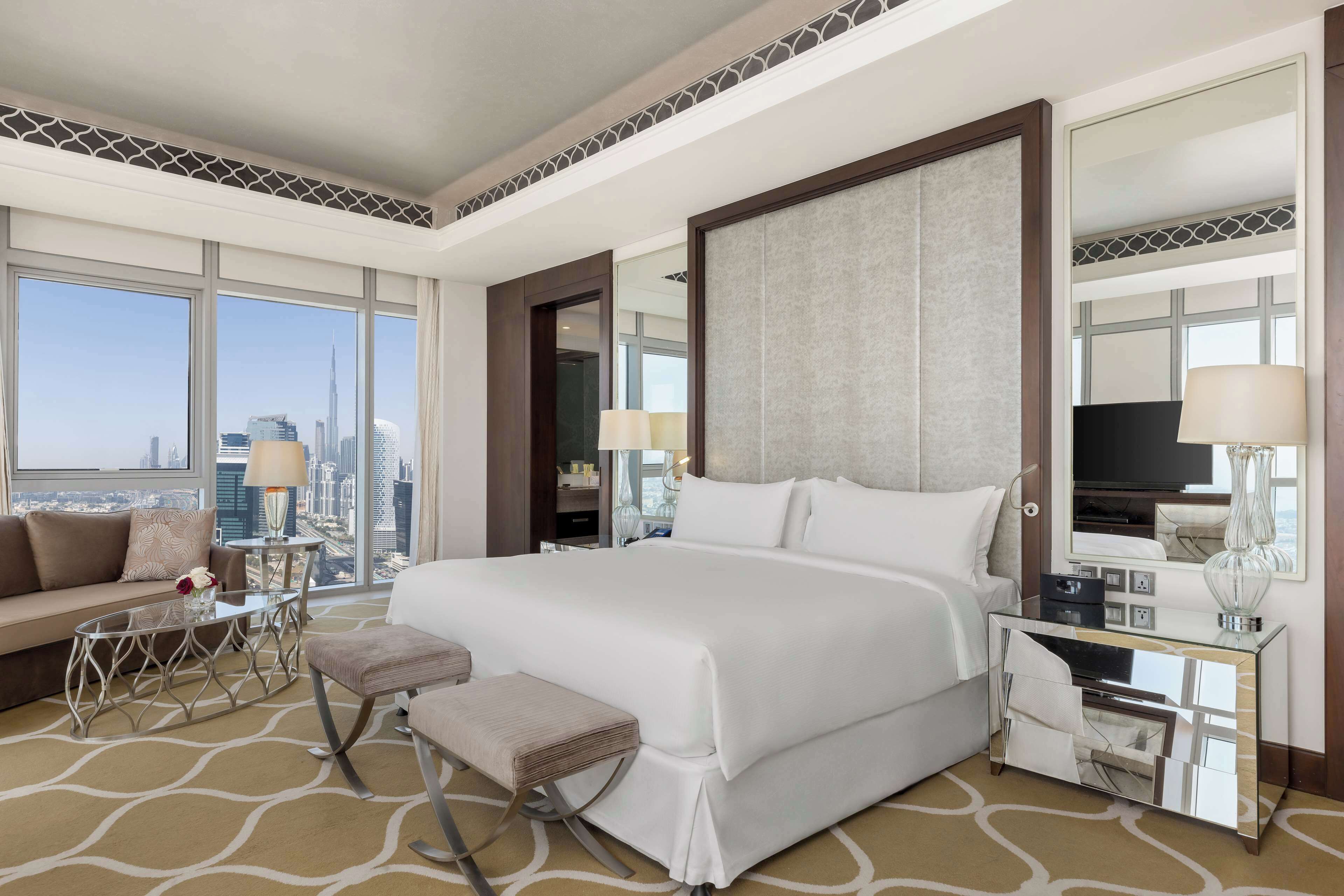 A modern hotel room with a large bed, a sitting area with a sofa and coffee table, a small table with lamps, and a large mirror.