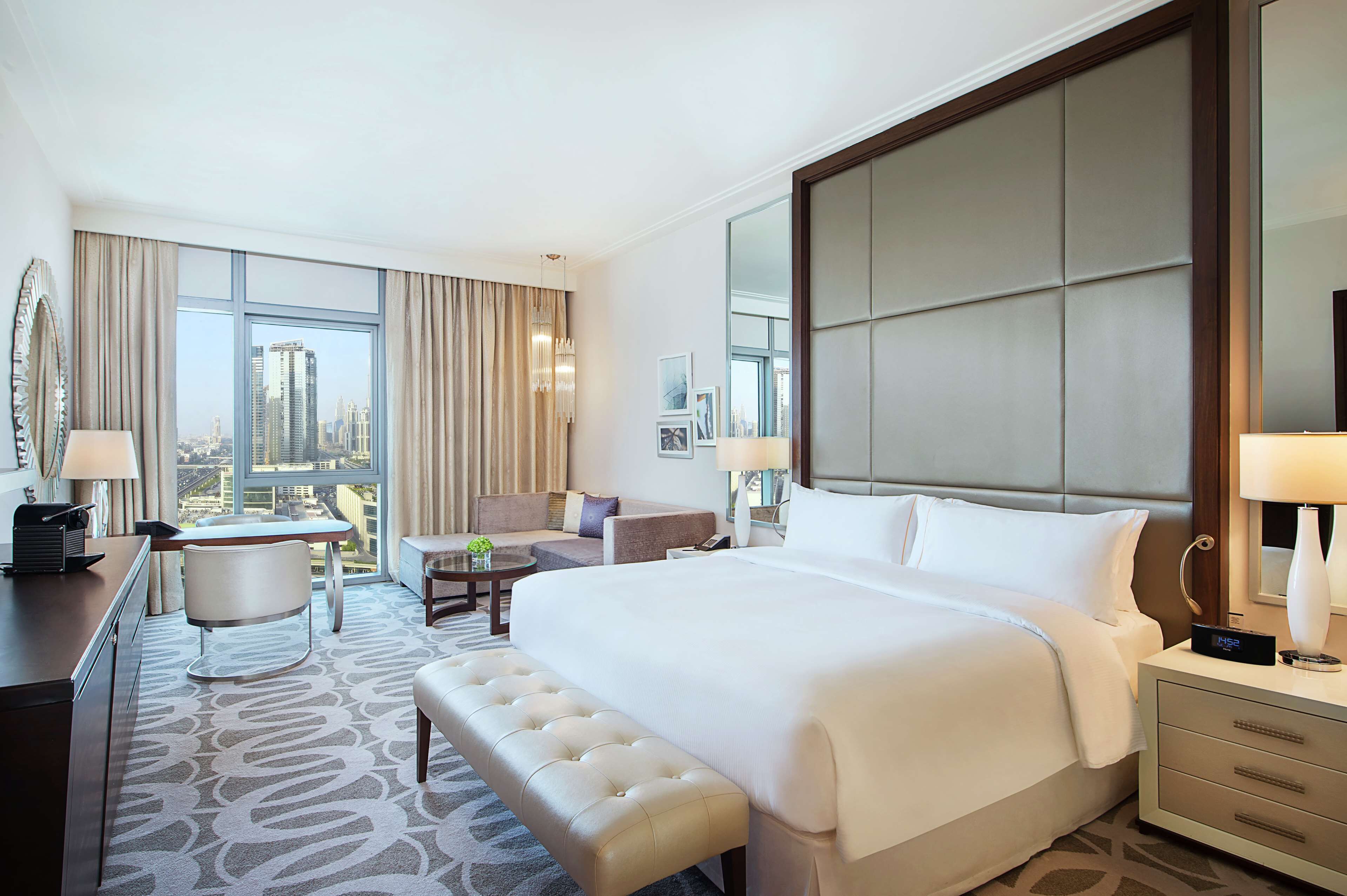 A hotel room with a large bed, a desk, a chair, a sofa, a bench, and a view of the city.