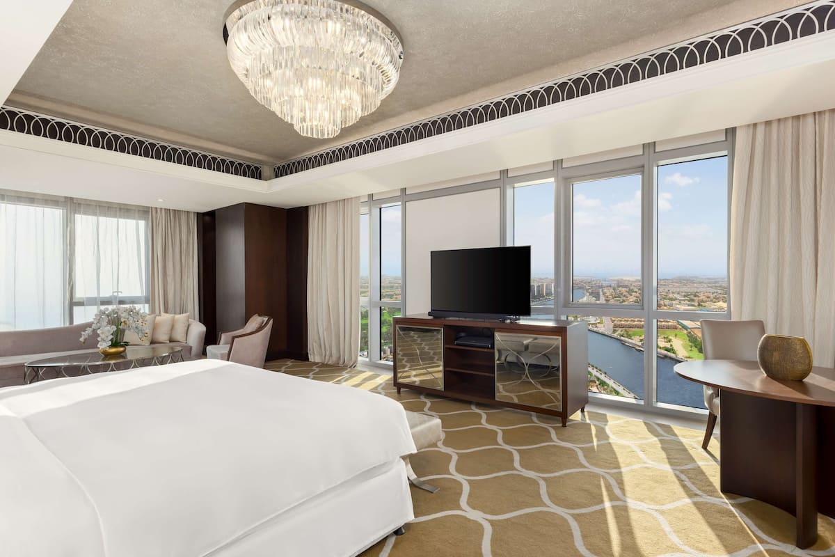 Habitación de hotel con una cama grande, televisor, balcón con vistas a un río y un candelabro.