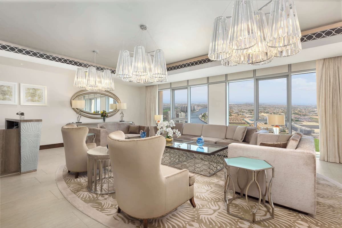 Amplia sala de estar con un sofá grande, dos sillones, una mesa de centro y un espejo decorativo. Varias lámparas colgantes y un ventanal con vistas a la ciudad.