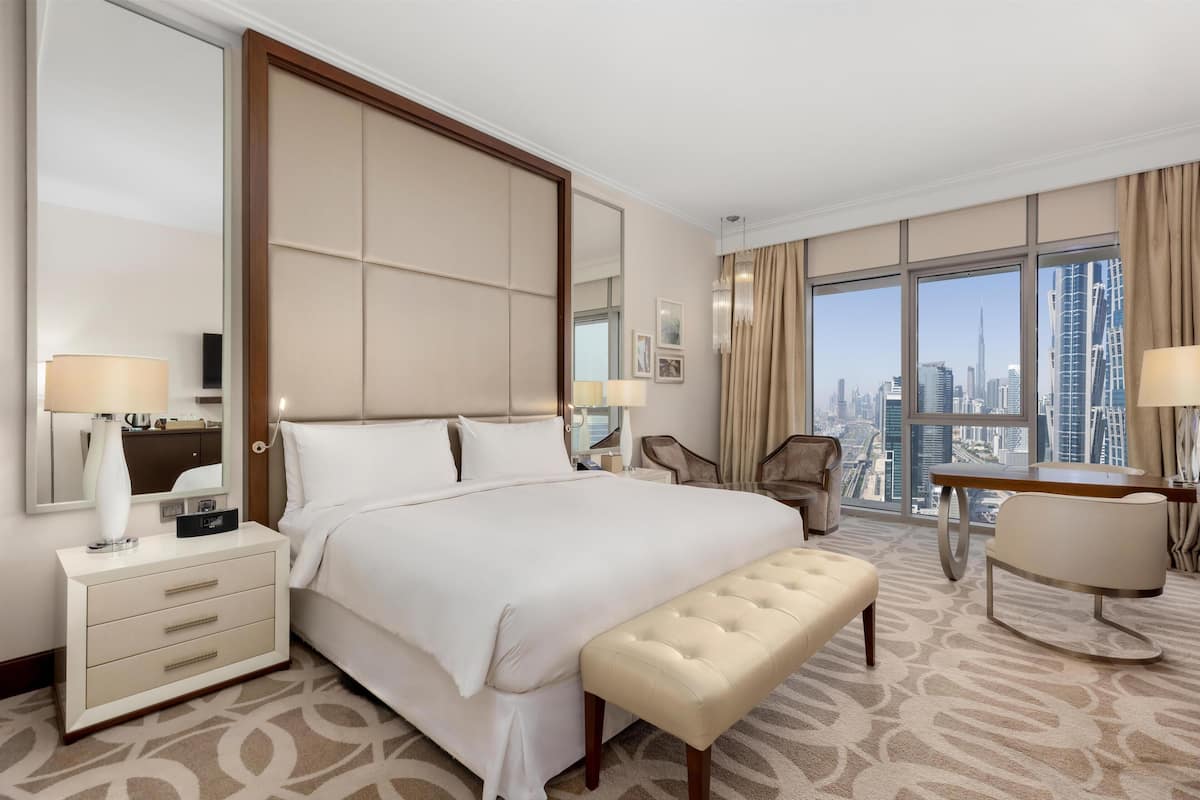 Una habitación de hotel moderna con una cama grande, una zona de estar con escritorio y vistas a la ciudad a través de amplias ventanas.