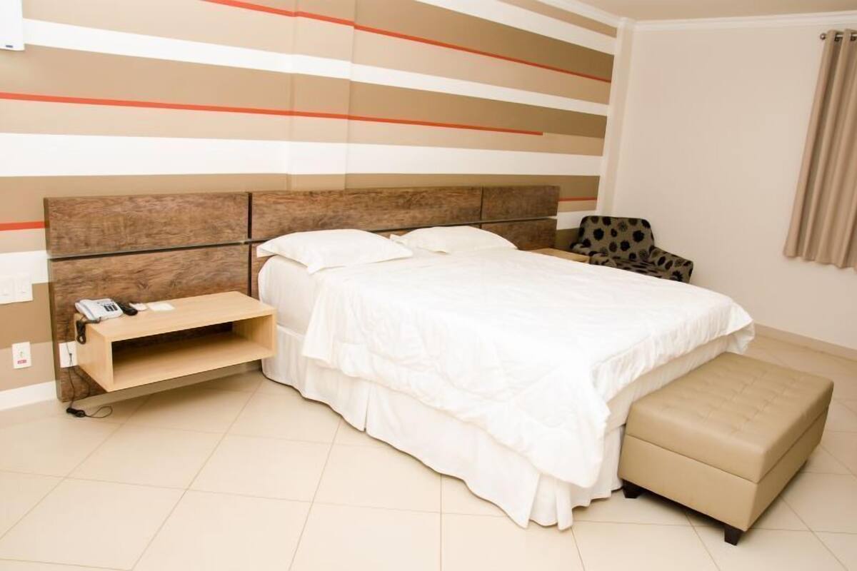 A bedroom with a bed, a bedside table with a phone, a chair, and a small ottoman.
