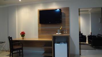 Una sala moderna con un televisor de pantalla plana montado en una pared de madera, una mesita con una botella de champán y copas, una silla y un refrigerador compacto.