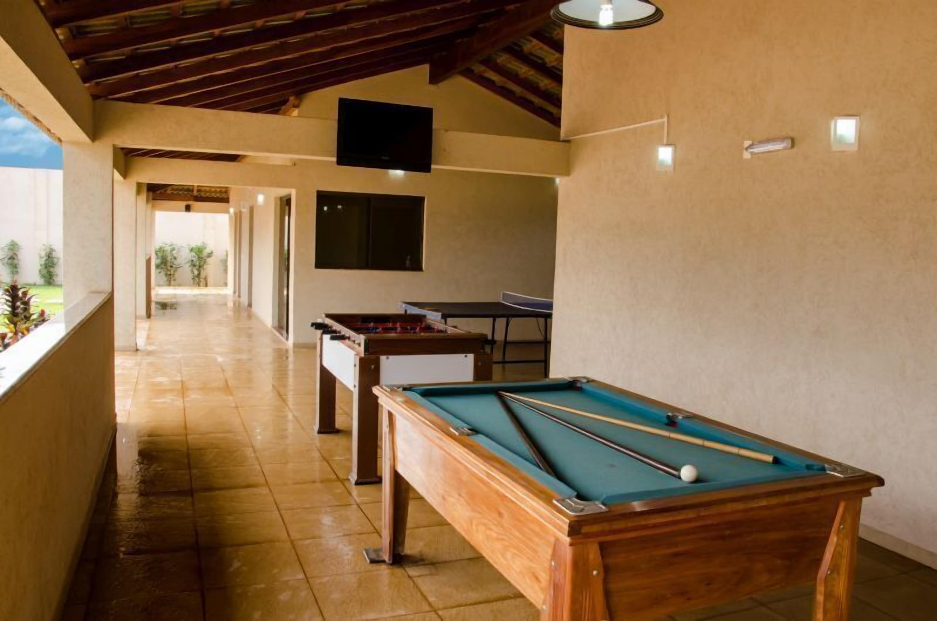 A pool table, a foosball table, and a ping pong table in a room with a high ceiling and a TV mounted on the wall.