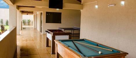 A pool table, a foosball table, and a ping pong table in a room with a high ceiling and a TV mounted on the wall.