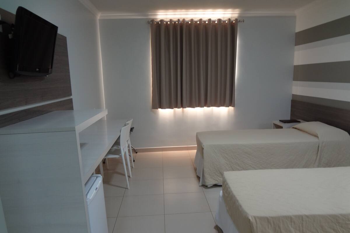 A hotel room with a bed, a desk, a chair, a TV, and a window with curtains.