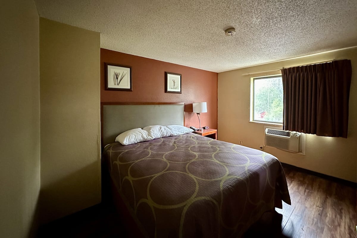 Ein Hotelzimmer mit Bett, Nachttisch, Lampe, Fenster mit Vorhängen und zwei gerahmten Bildern an der Wand.