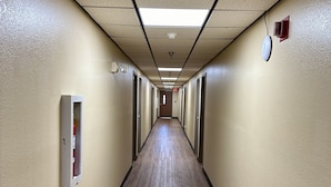 A long, narrow hallway with wooden flooring and beige walls, featuring fire safety equipment and emergency exit signs. - American Inn (Alexander City)