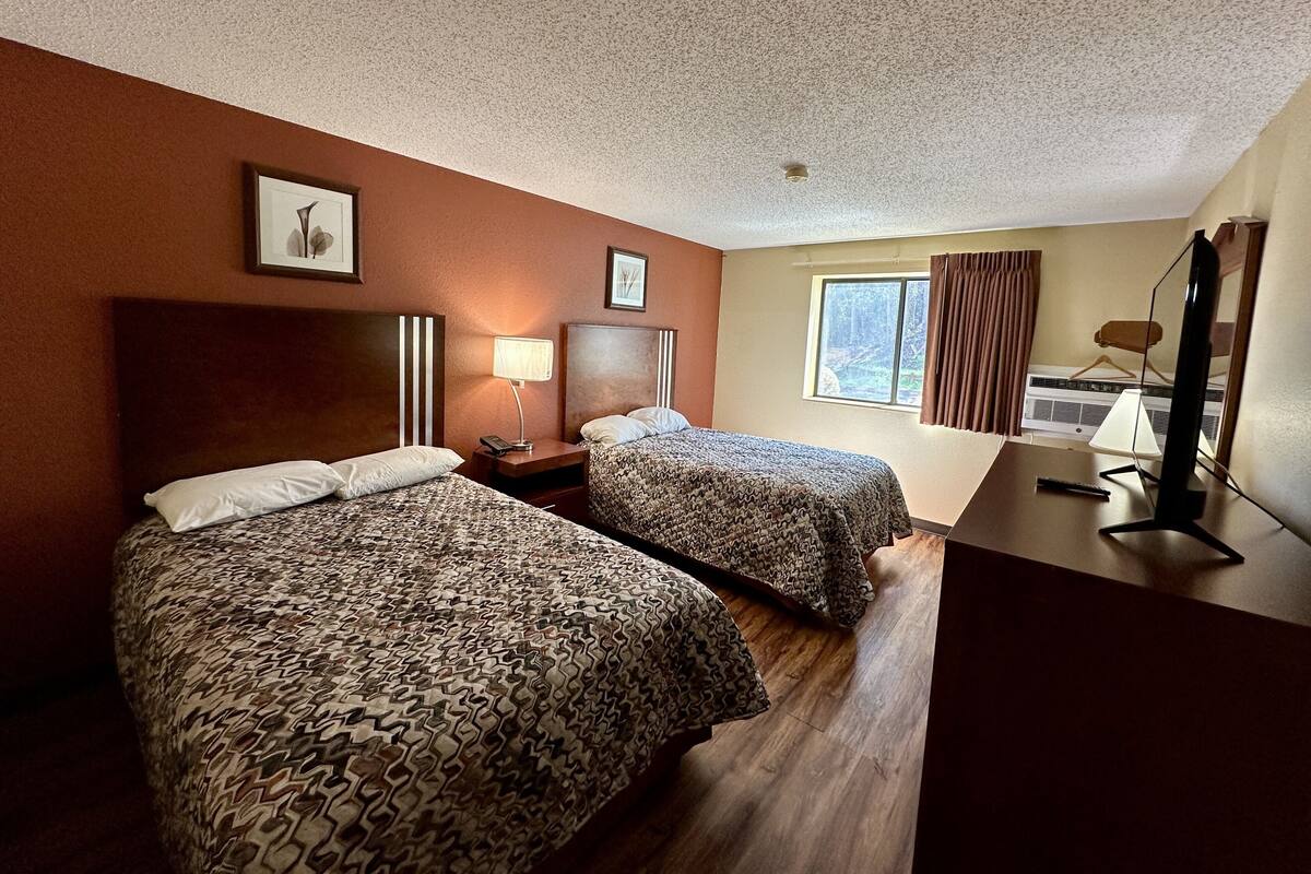 Ein Hotelzimmer mit zwei Betten, einem Schreibtisch mit Computer, einer Lampe und einem Fenster mit Vorhängen.