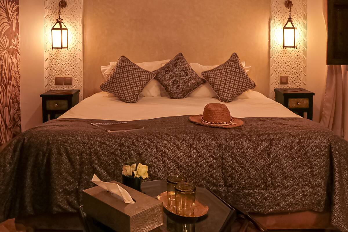 A neatly made bed with patterned pillows, a hat, a tissue box, and a tray with jars on a bedside table.