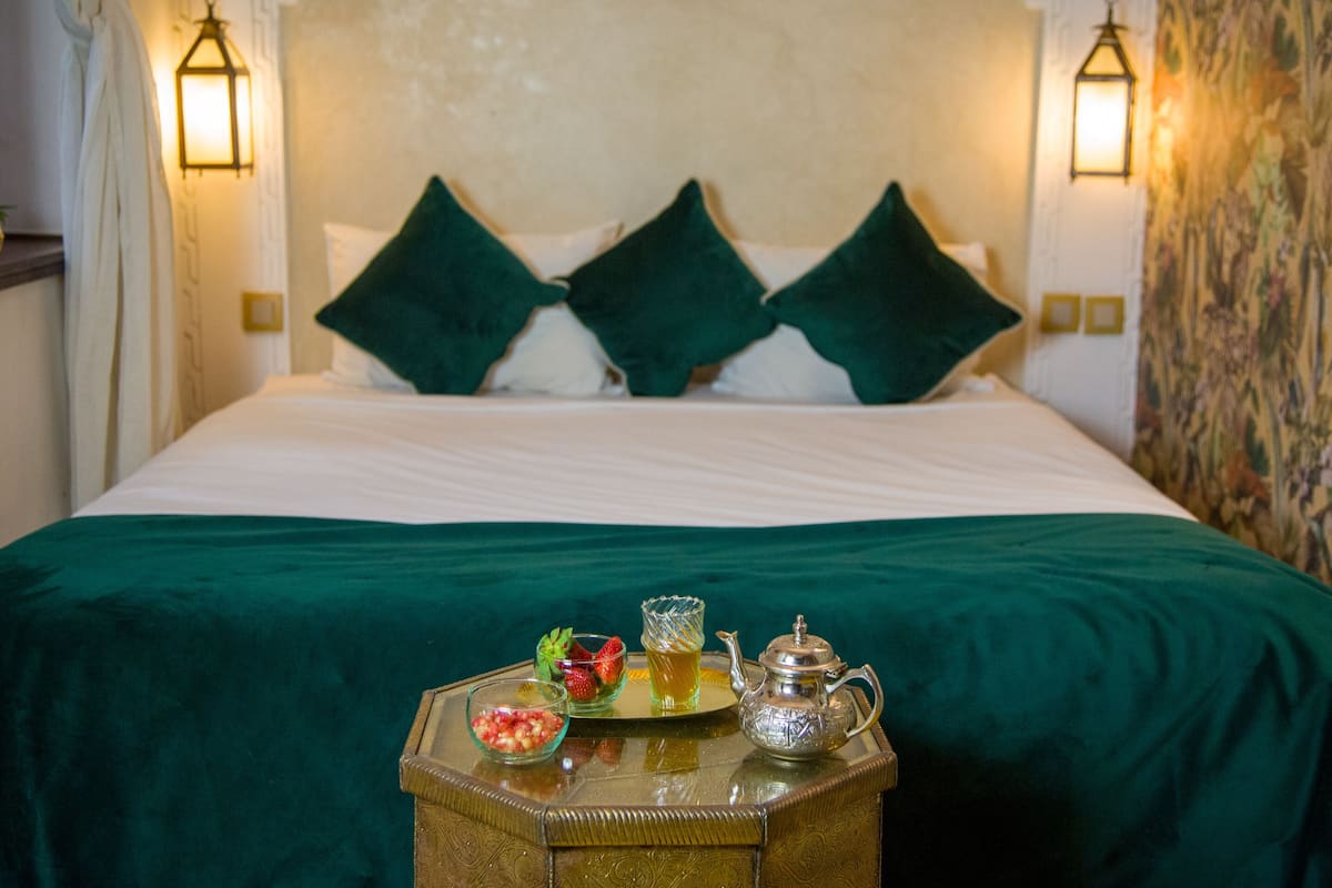 A neatly made bed with green pillows and a tray with a teapot, a glass of tea, and a bowl of fruit on the side.