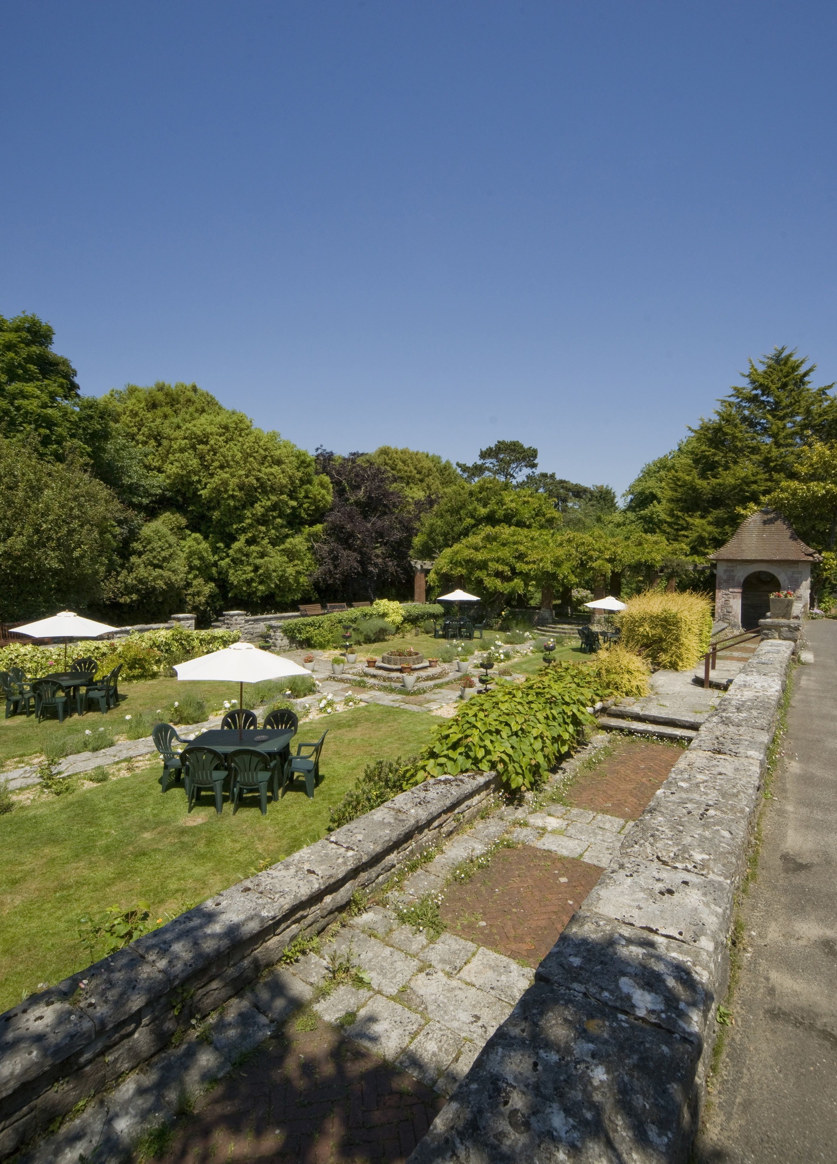 Un giardino esterno con sentieri in pietra, prati verdi e diversi tavoli sotto ombrelloni.