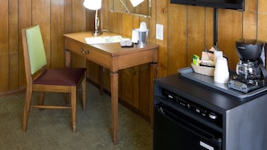 A room with wooden panel walls, a desk with a lamp, a chair, a television, and a coffee maker. - Sunset Motel (Moriarty)